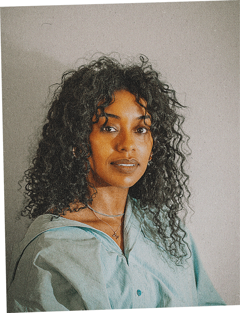 Headshot of a woman with long curly black hair smiles slightly wearing a loose light blue buttoned shirt. 