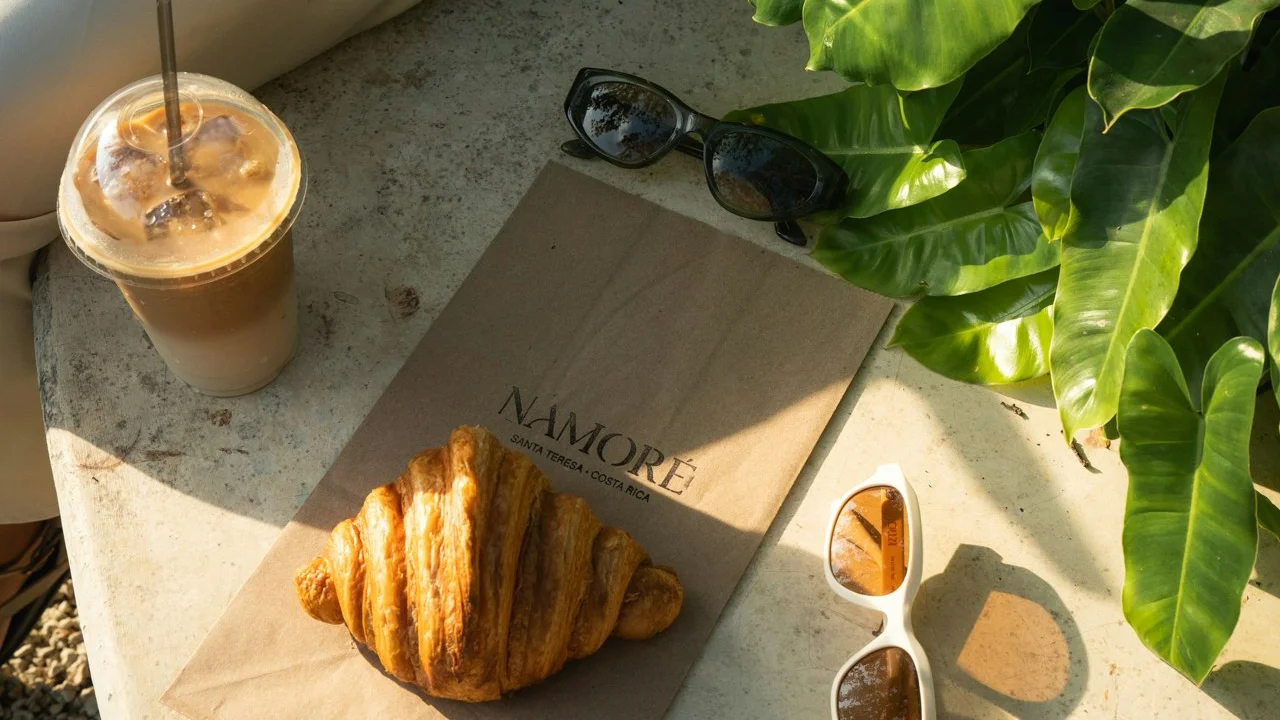 Iced coffee and a croissant on a café table with sunglasses and tropical leaves