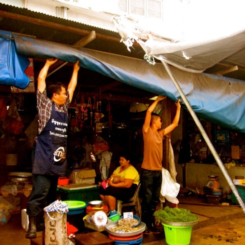 두 사람이 야외 시장 가판대 위에 파란 방수포를 조정하고 있고, 앉아 있는 여자가 그것을 지켜보고 있습니다. 다양한 상품들이 진열되어 있습니다.