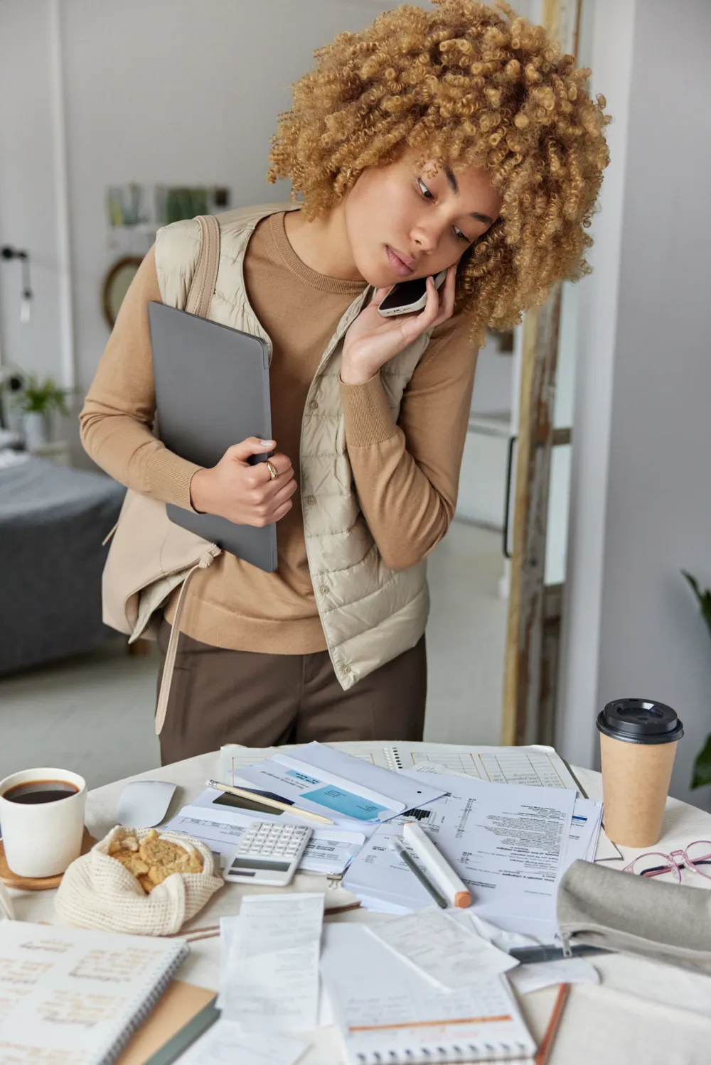 Woman stood in front of messy desk on the phone holding a laptop