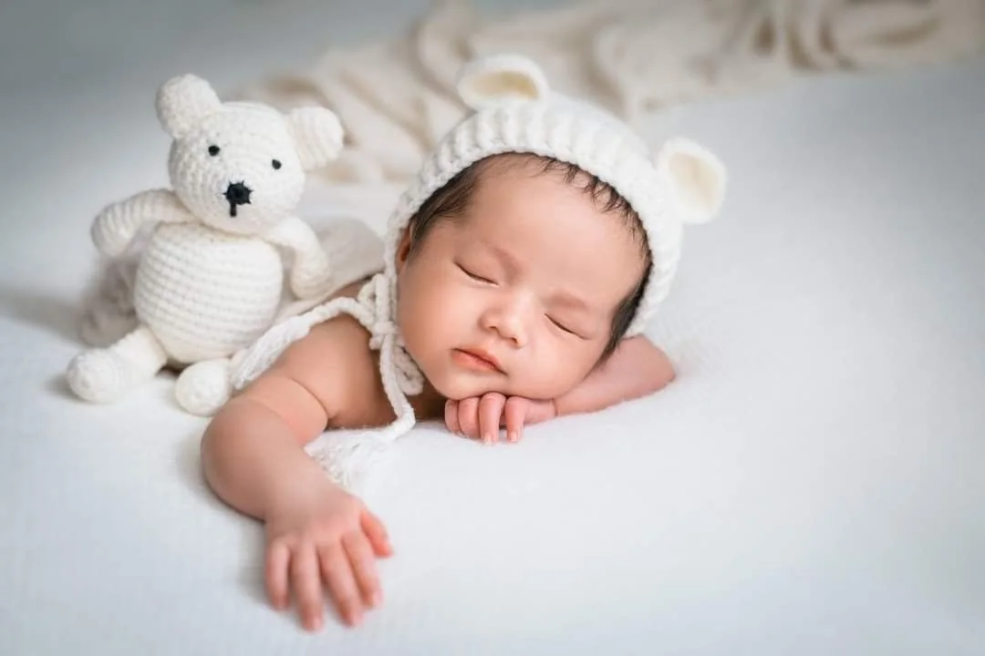Sleeping baby in a white bear bonnet beside a knitted teddy