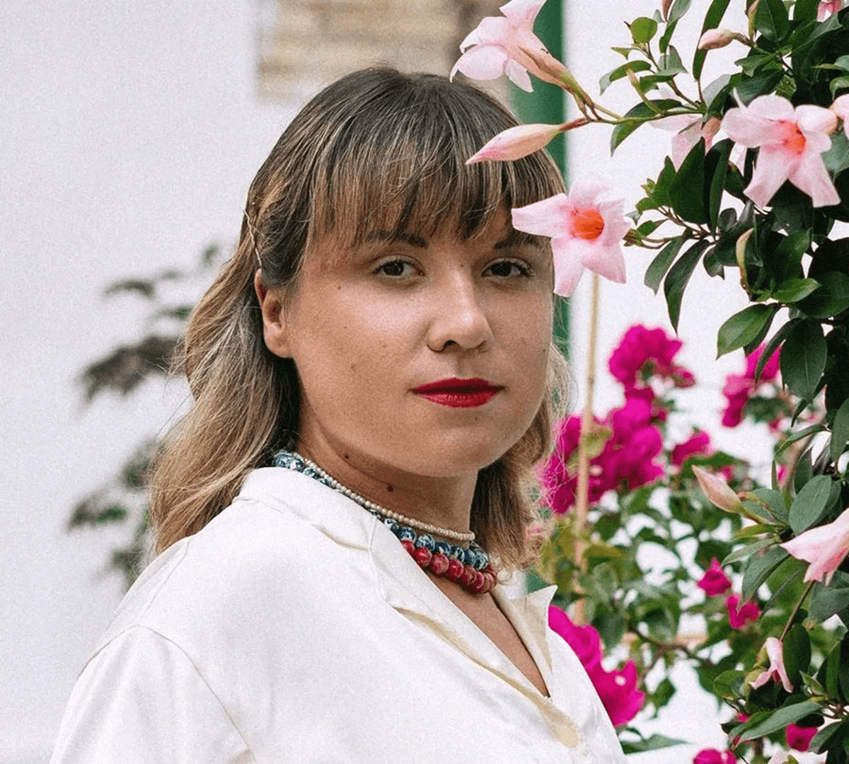 A person with light hair gazes at the camera while standing next to a lush flowering plant featuring pink and magenta blooms.