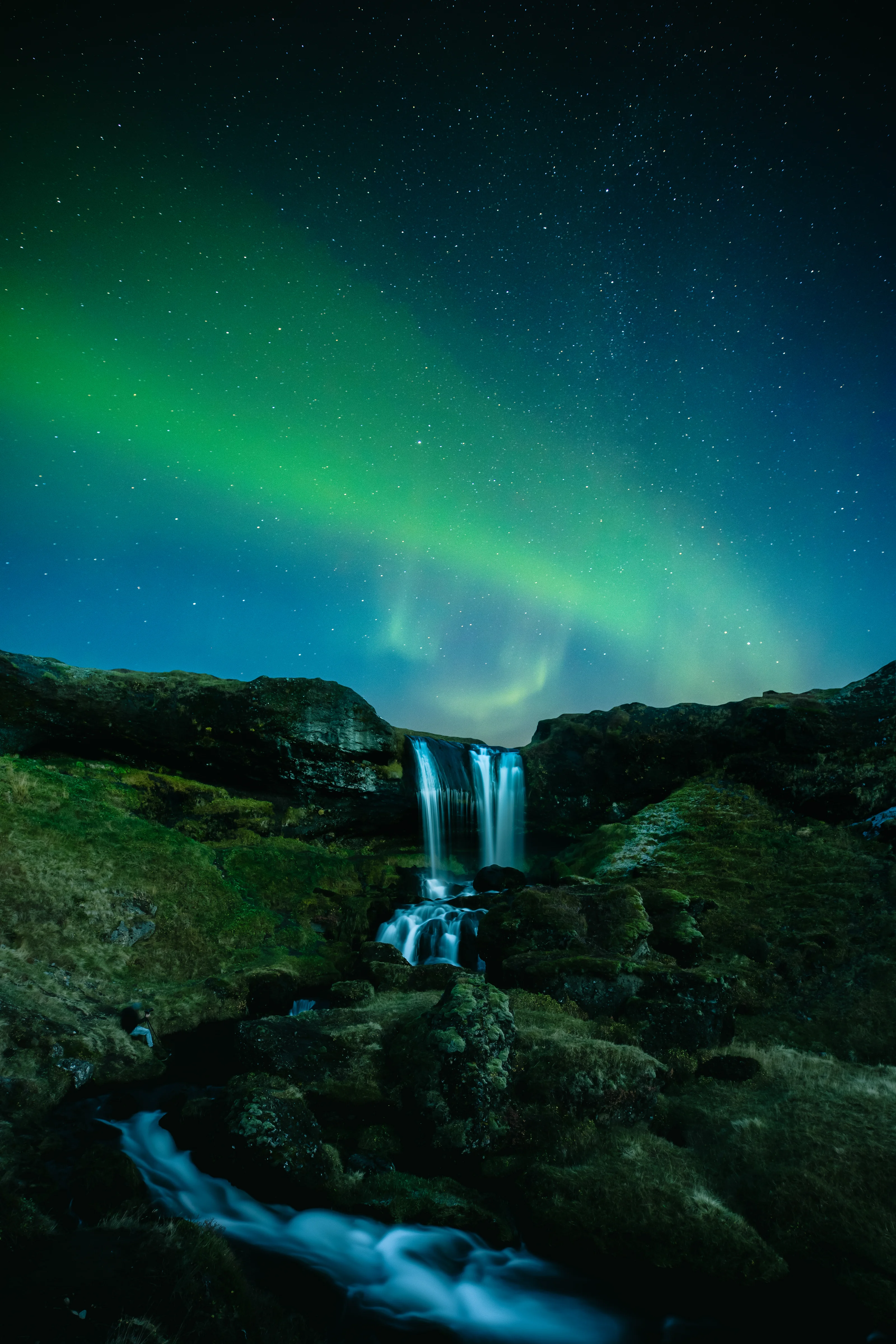 Vibrant green Northern Lights dancing over a tiered waterfall in Iceland at night. Photo by Witness.
