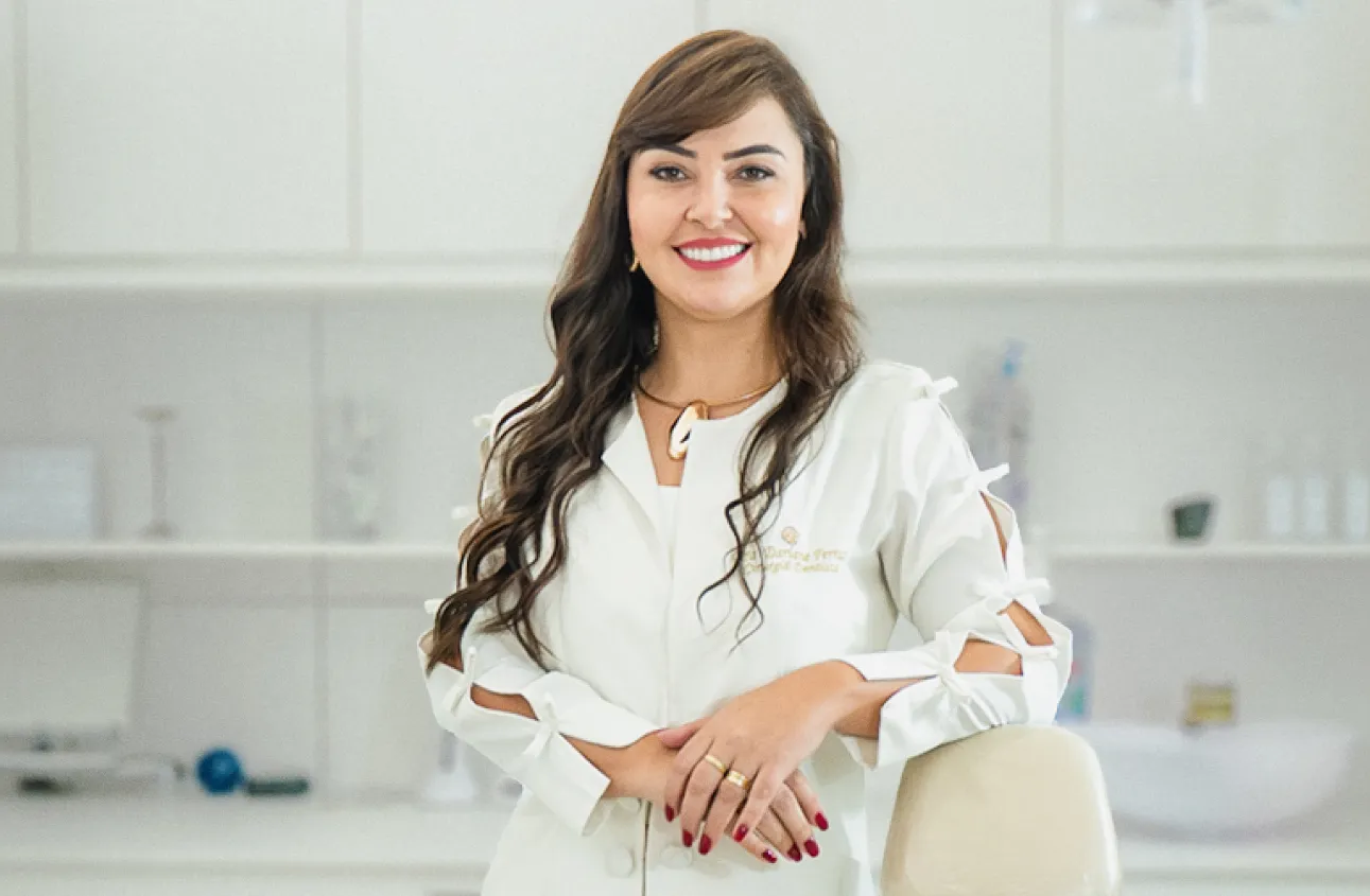 Retrato de uma dentista, a Dra. Dariane sorridente em um consultório odontológico. Ela está em pé, usando jaleco branco e apoiando as mãos sobre uma cadeira odontológica. Ao fundo aparecem equipamentos e pia do consultório em um ambiente claro e organizado.