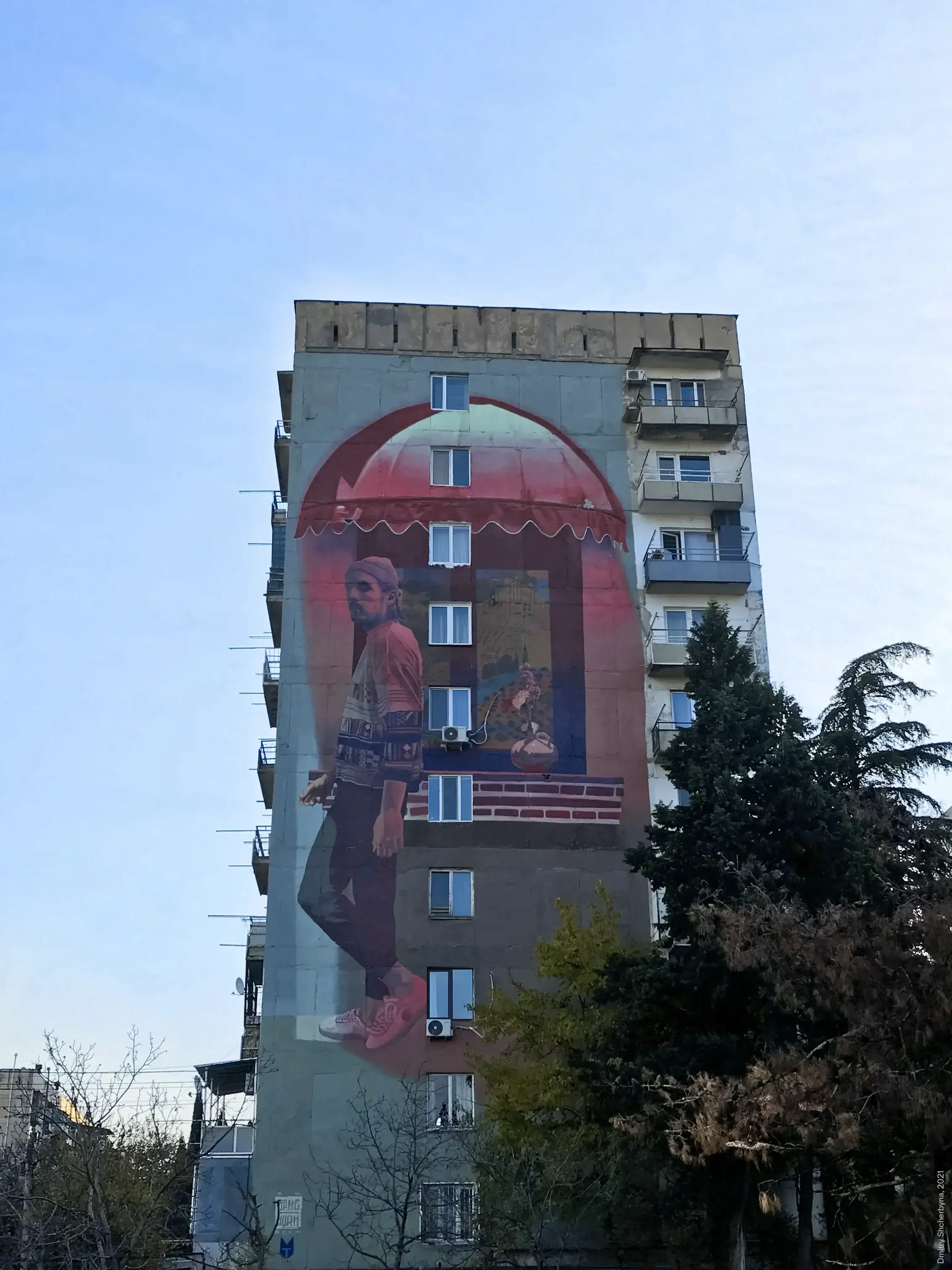 A tall mural on a Soviet-era apartment building shows a young man in a patterned sweater and beanie walking inside, contrasting the warm interior with the grey exterior of the building.