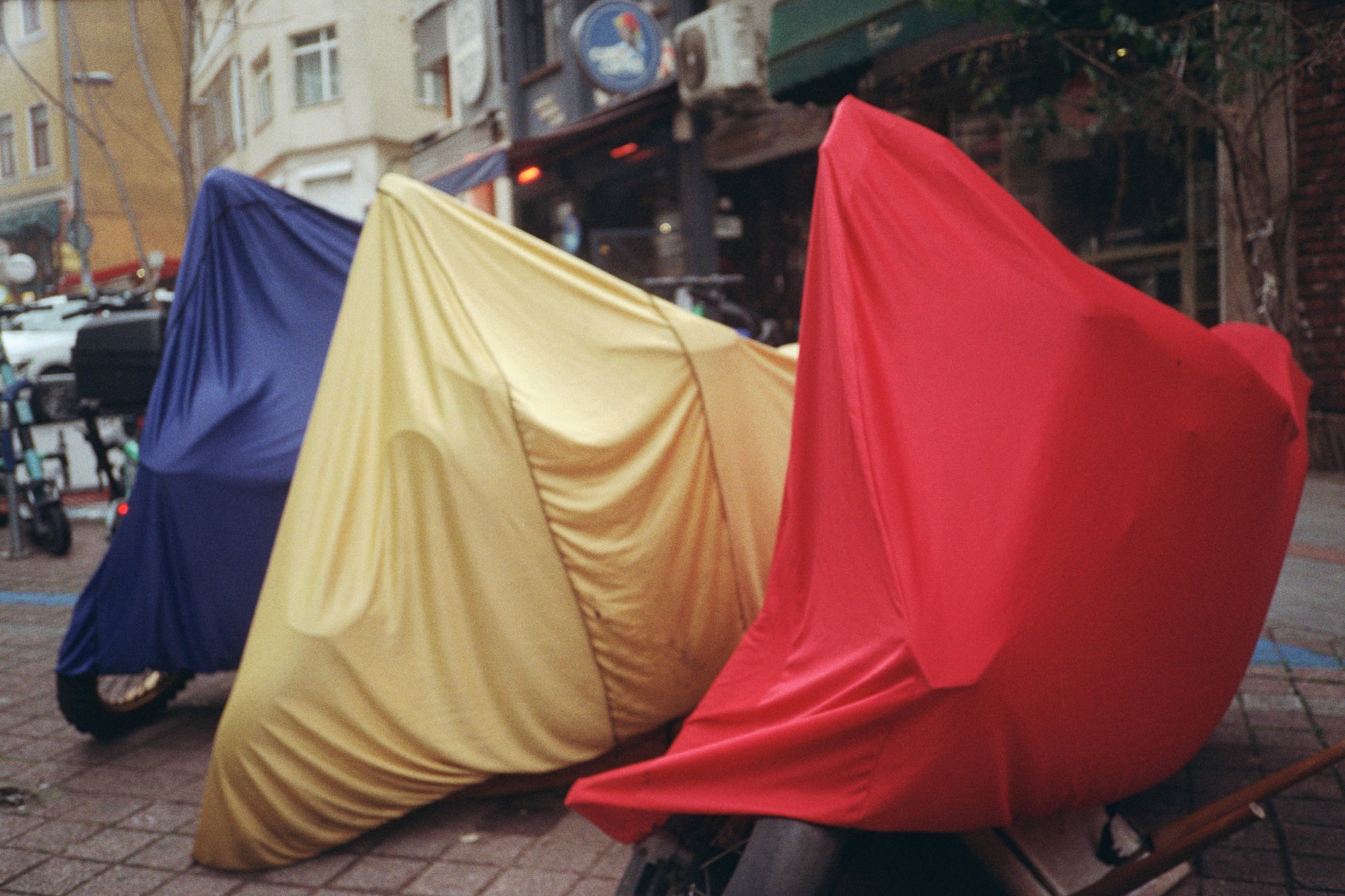 Three motorcycles covered with vibrant blue, yellow, and red fabric covers are parked on a cobblestone street in an urban setting with buildings in the background.