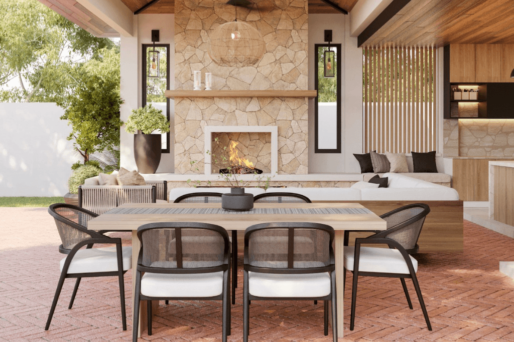 Open-air dining and living space with wood furniture, stone fireplace, vaulted ceiling, and landscaped surroundings.