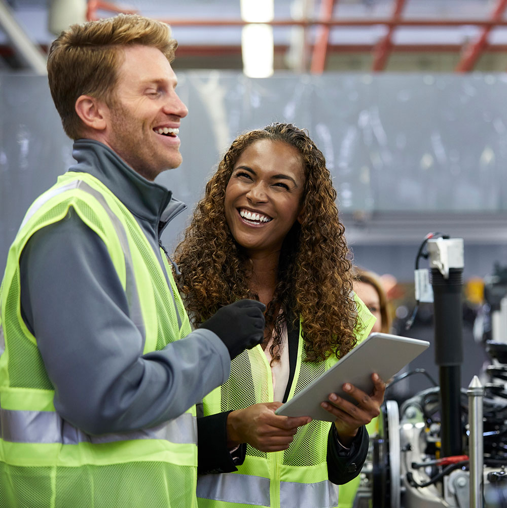 Two industrial workers wearing high-visibility safety vests share a laugh in a manufacturing facility. A woman with curly hair holds a tablet while standing next to a smiling male colleague. Industrial equipment and machinery are visible in the background, suggesting a factory or warehouse environment where safety and teamwork are emphasized.