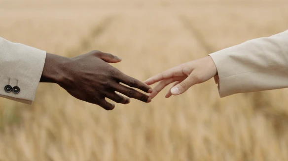 A close-up shot of two hands, one dark-skinned and one light-skinned, reaching out to touch against a blurred background of a golden field.