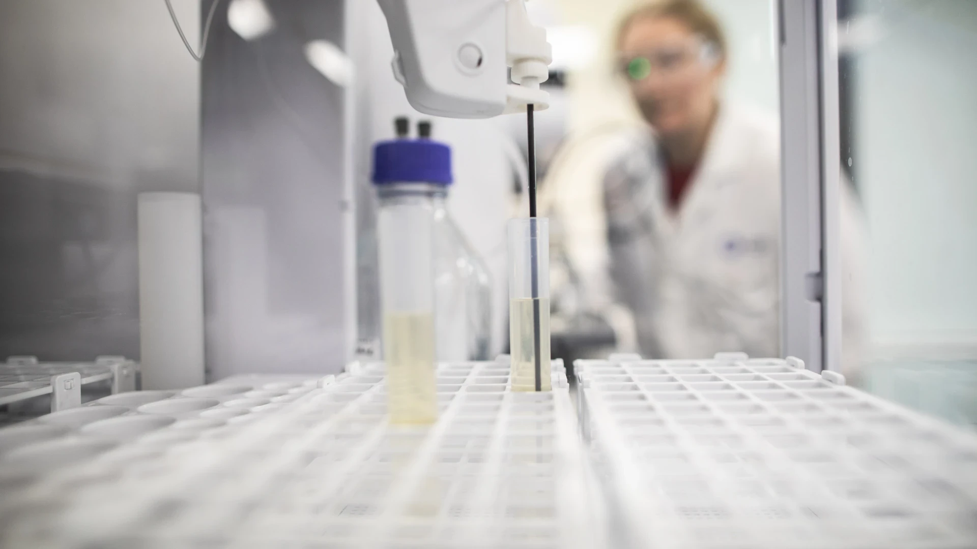 Automated sampler probe dipping into a sample tube in a rack, with a CPI scientist watching behind.