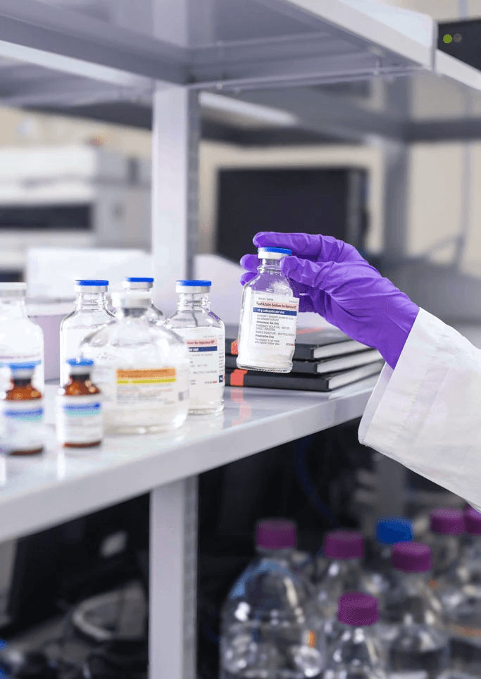 Gloved hand holding a small glass vial on a laboratory shelf with other vials and bottles.