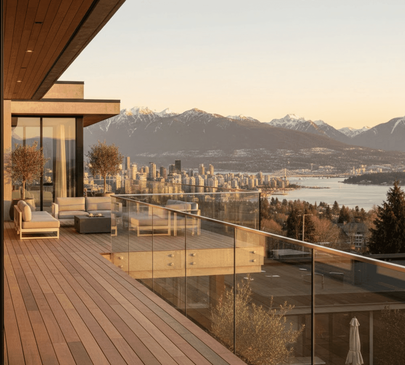 Luxury home deck with transparent frameless glass railings, panoramic view of Vancouver North Shore mountain range, snow-capped peaks, city skyline in middle distance, golden hour lighting, contemporary architecture, crystal clear glass panels, photorealistic
