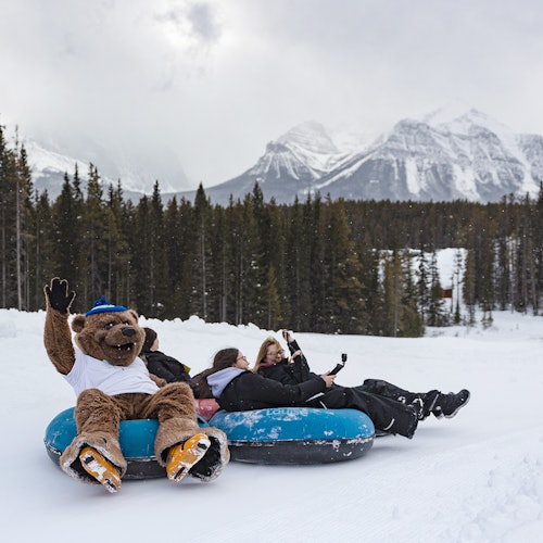 ผู้คนกำลังเล่นสโนว์ทูบ หนึ่งคนแต่งตัวเป็นมาสคอตหมี โดยมีภูเขาหิมะและต้นสนอยู่เบื้องหลัง