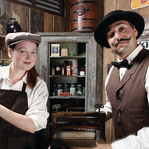 Two people in vintage attire stand in an old-fashioned shop with various goods on shelves behind them.