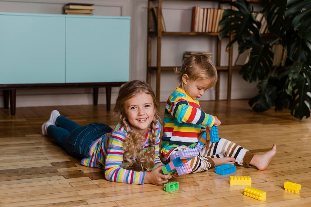 Two young children play together on a wooden floor, building with colorful toy blocks in a cozy living room. The image represents family life and the comfort of homeownership made possible through conventional home loans from Chris Lewis Home Loans.