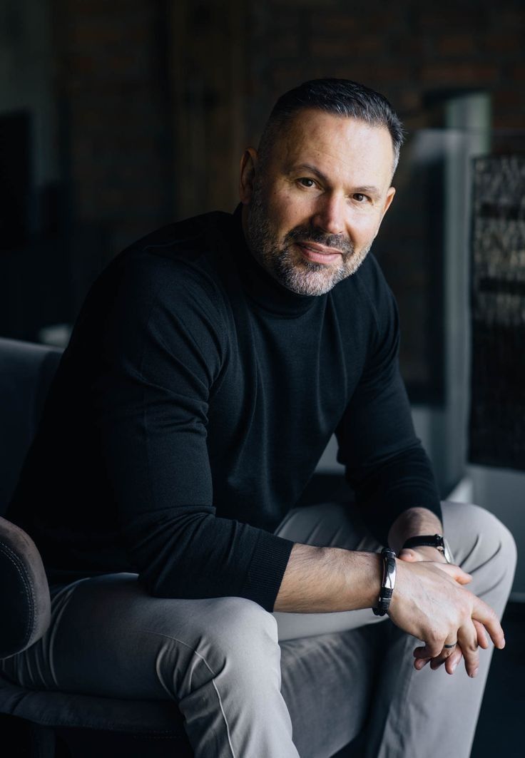 A man in a black turtleneck and gray pants sits casually, leaning forward with fingers intertwined. The mood is relaxed and contemplative.