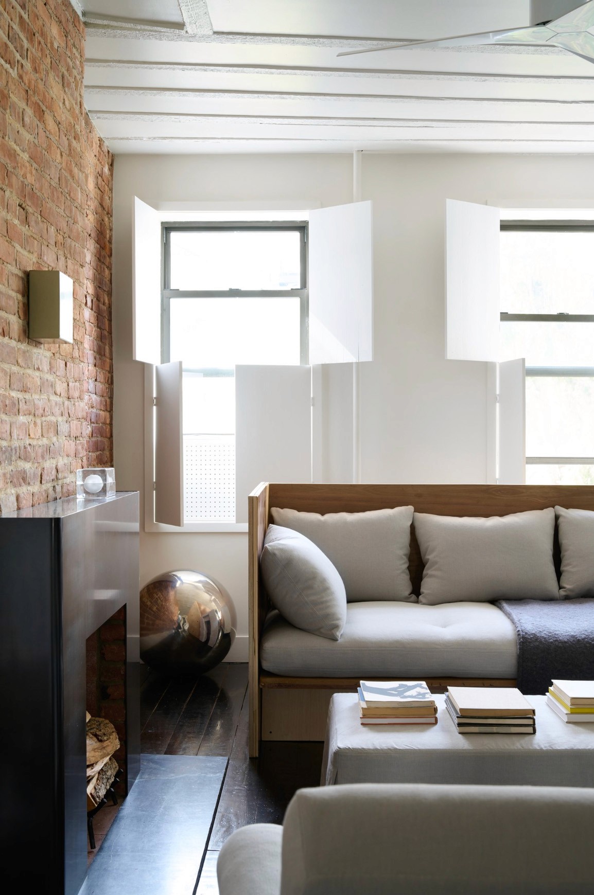 Living room with minimalist sofa, upholstered ottoman with books, and white wooden shutters.