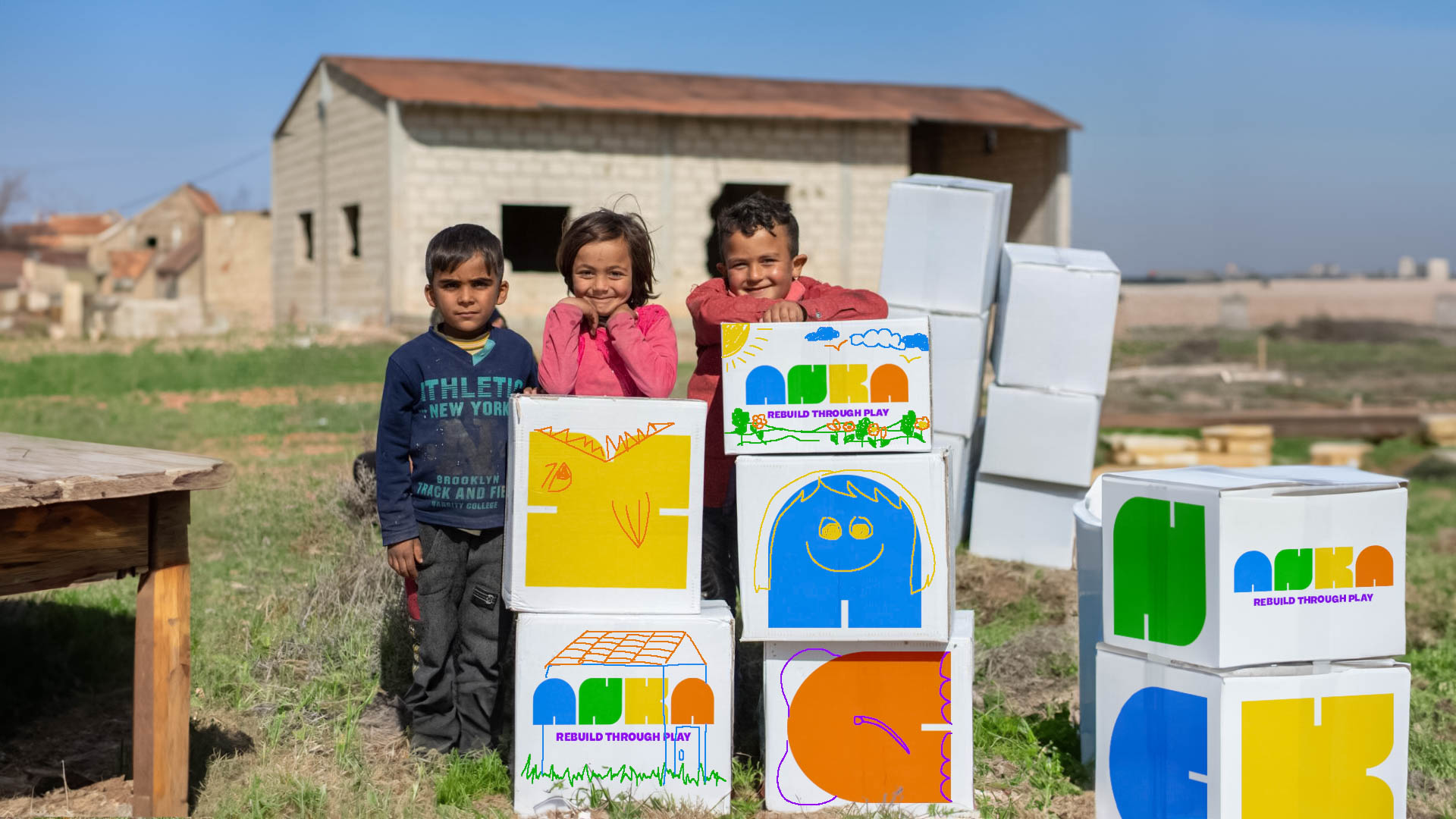 Three children stand in a grassy area in front of a light-colored building. The child on the left wears a dark blue long-sleeved shirt with "ATHLETIC NEW YORK" written on it. The middle child wears a pink jacket. The child on the right wears a red jacket. All three children are smiling and looking toward the camera. Behind them are several white cardboard boxes stacked, with colorful drawings and text on them, including “REBUILD THROUGH PLAY.” A wooden table is visible to the left. The background includes more buildings and a clear sky.