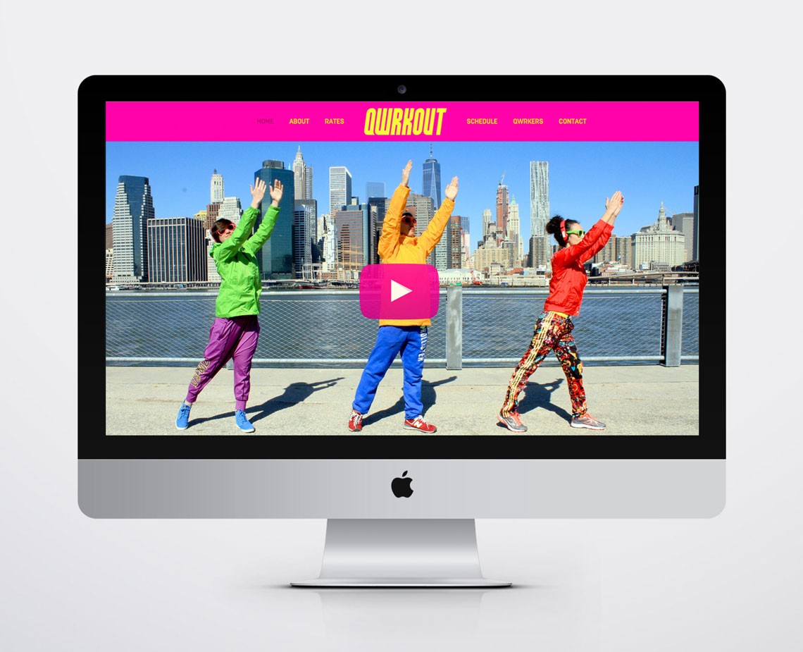 A silver Apple iMac computer displays a webpage titled “QUIRKOUT” with a pink banner at the top. Below the title, three figures are shown in a dynamic pose: one in green, one in yellow, and one in red. The central figure’s right arm is raised, and the left arm is raised in a “V” shape. The background features a cityscape with tall buildings under a clear blue sky. A pink play button is centered on the page. The webpage includes navigation links: “HOME,” “RATES,” “SERVICES,” “QUIRKERS,” and “CONTACT.”