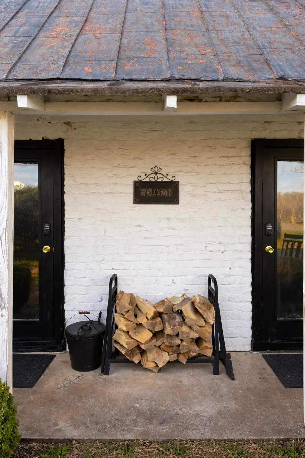 Stacked firewood and 'Welcome' sign on a white brick porch.