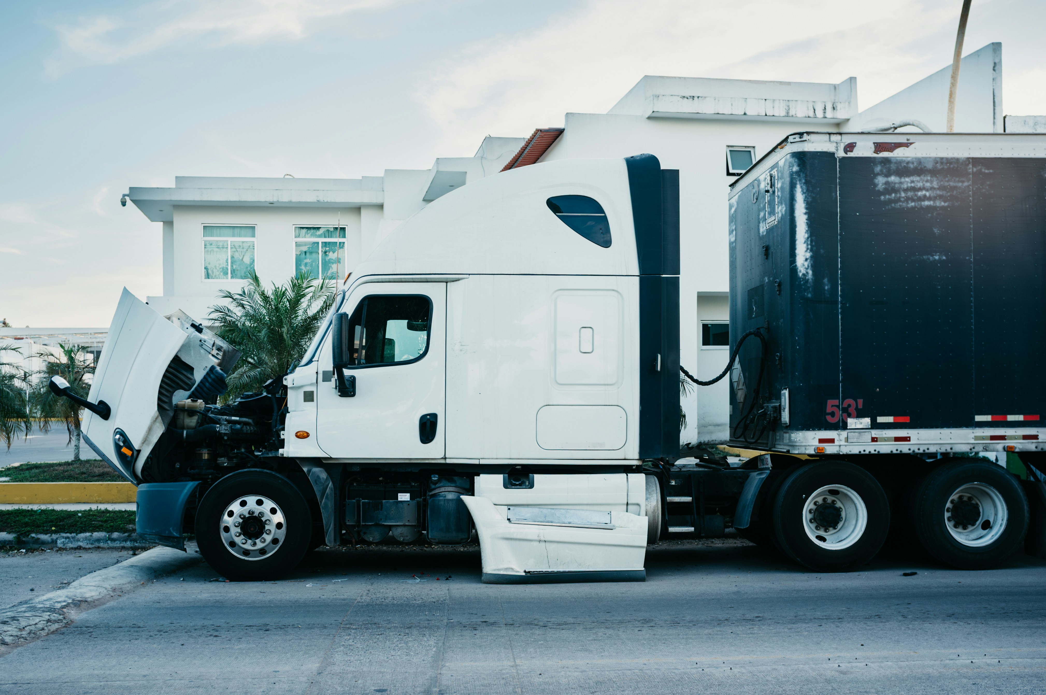 A white semi-truck with its hood open for engine inspection, coupled to a 53-foot dark trailer, parked outside a commercial facility — illustrating commercial vehicle maintenance and FMCSA compliance.