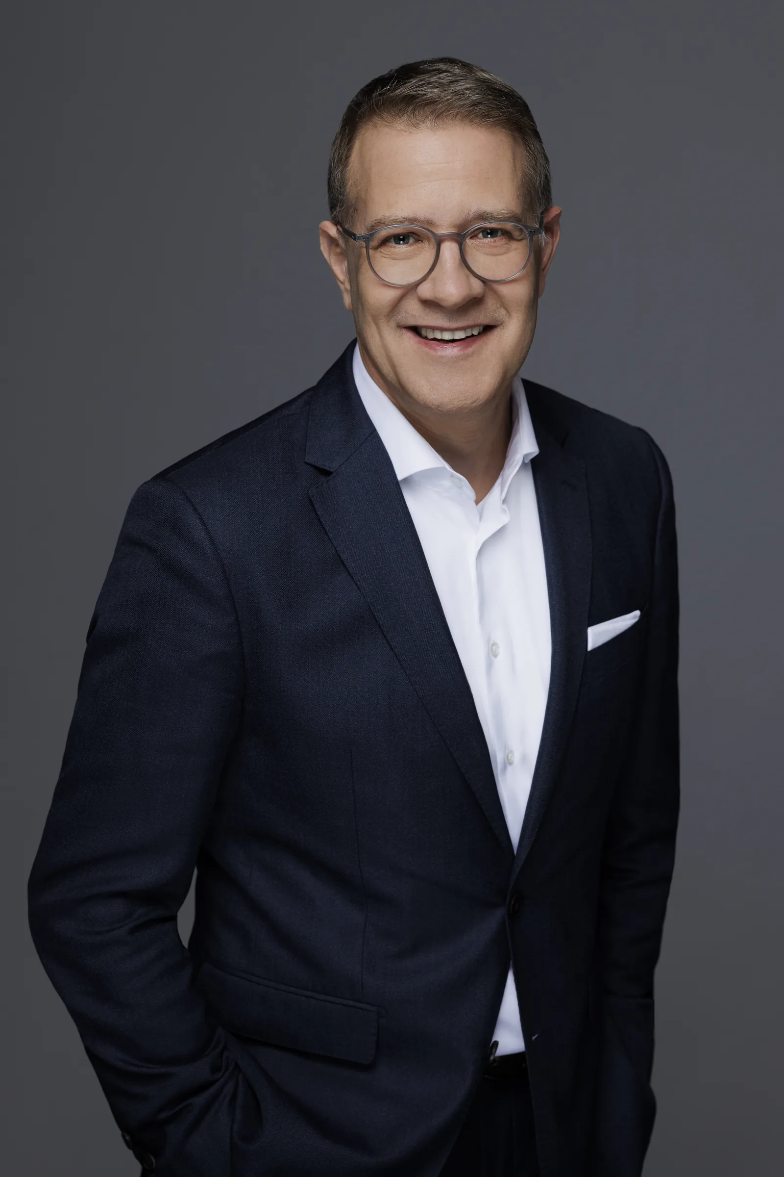 Modernes, lockeres Studio-Porträt von Adrian Grasse im dunklen Anzug mit offenem Hemdkragen vor grauem Hintergrund.