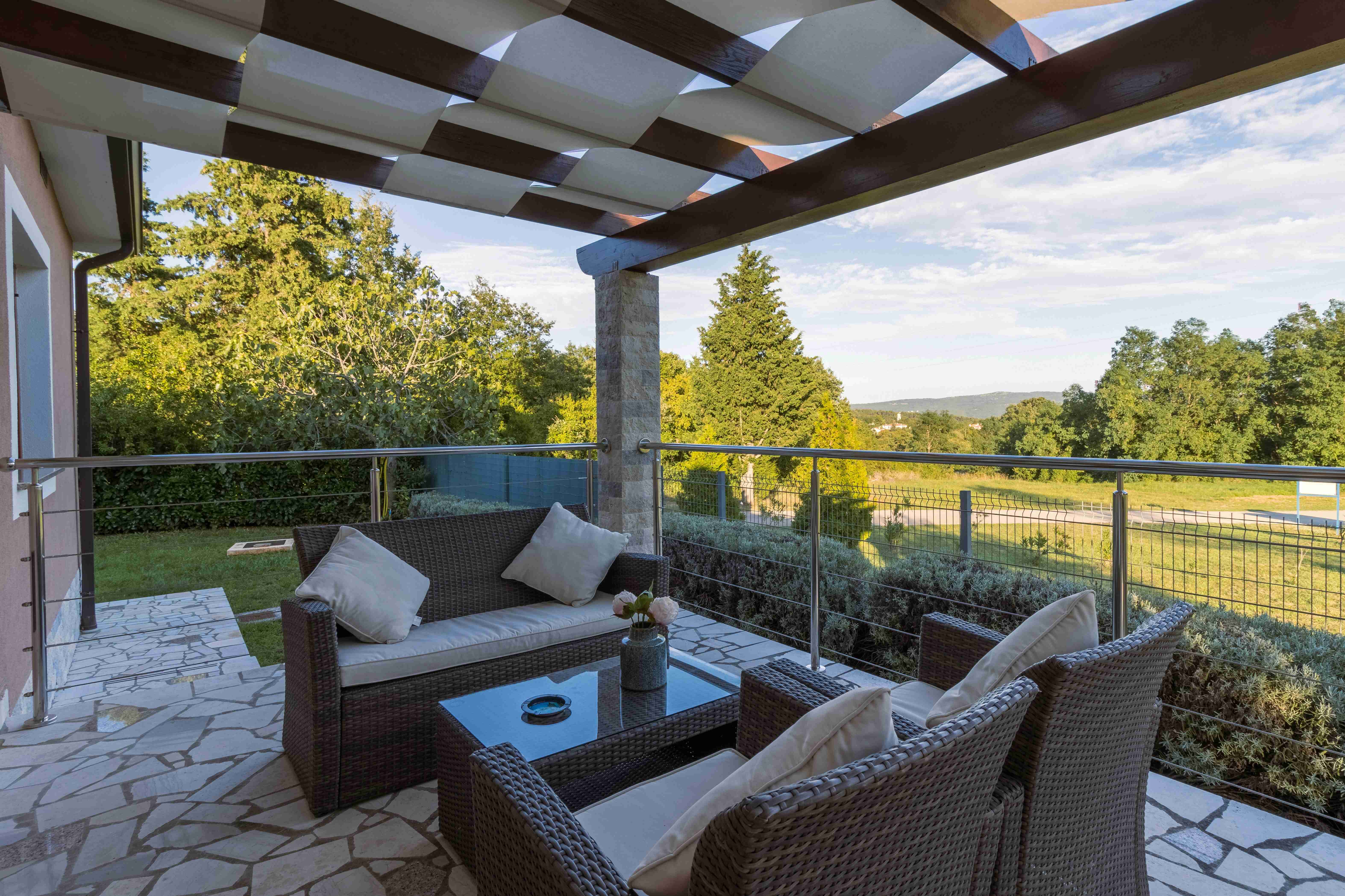 Outdoor patio seating area with wicker furniture, stone flooring, and a pergola overlooking trees and open views.