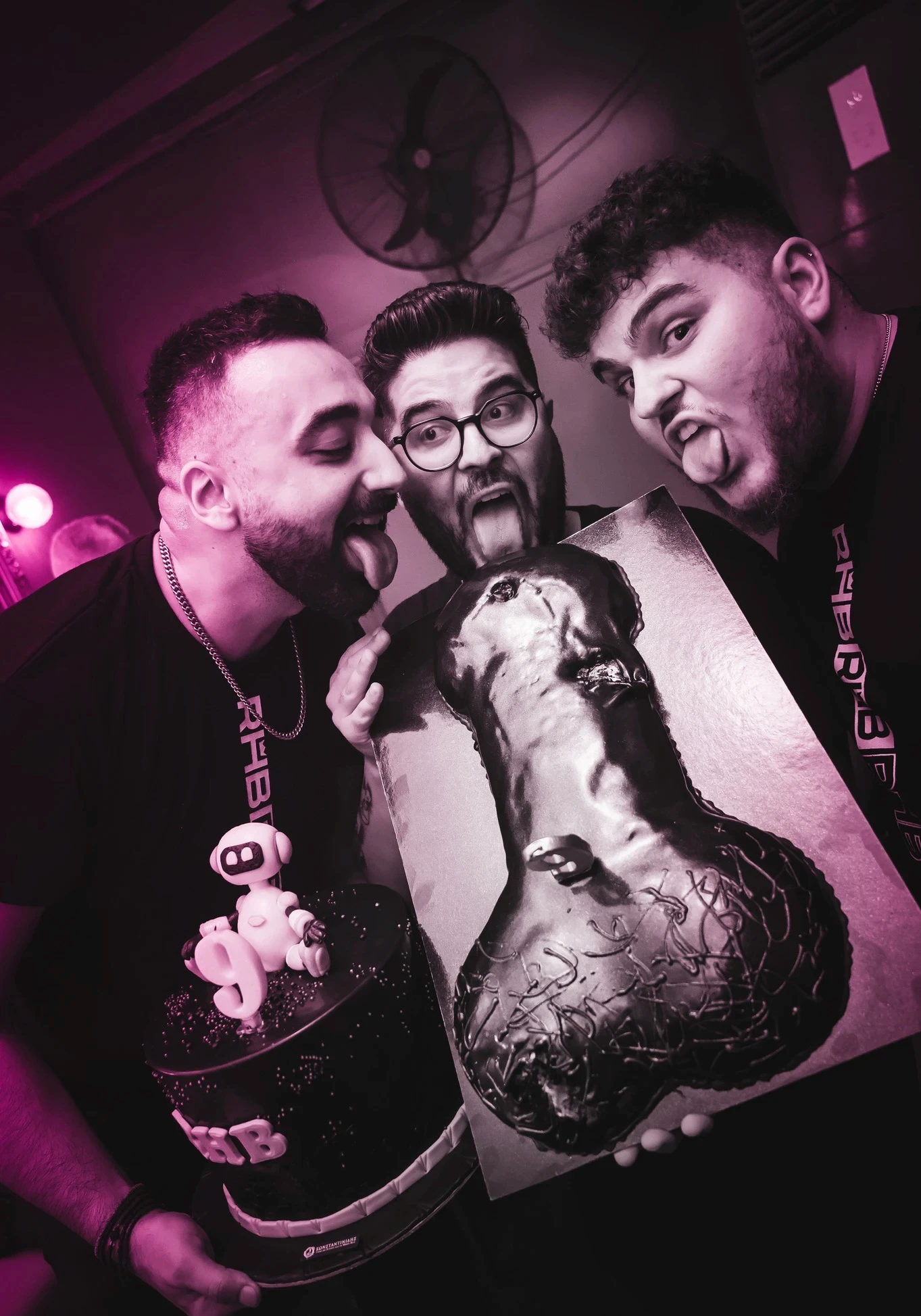 Three men make silly faces, sticking their tongues out as they pose with two cakes. One man holds a round black cake with an "RHB" logo and a topper, while the other two hold a large, phallic-shaped chocolate cake.