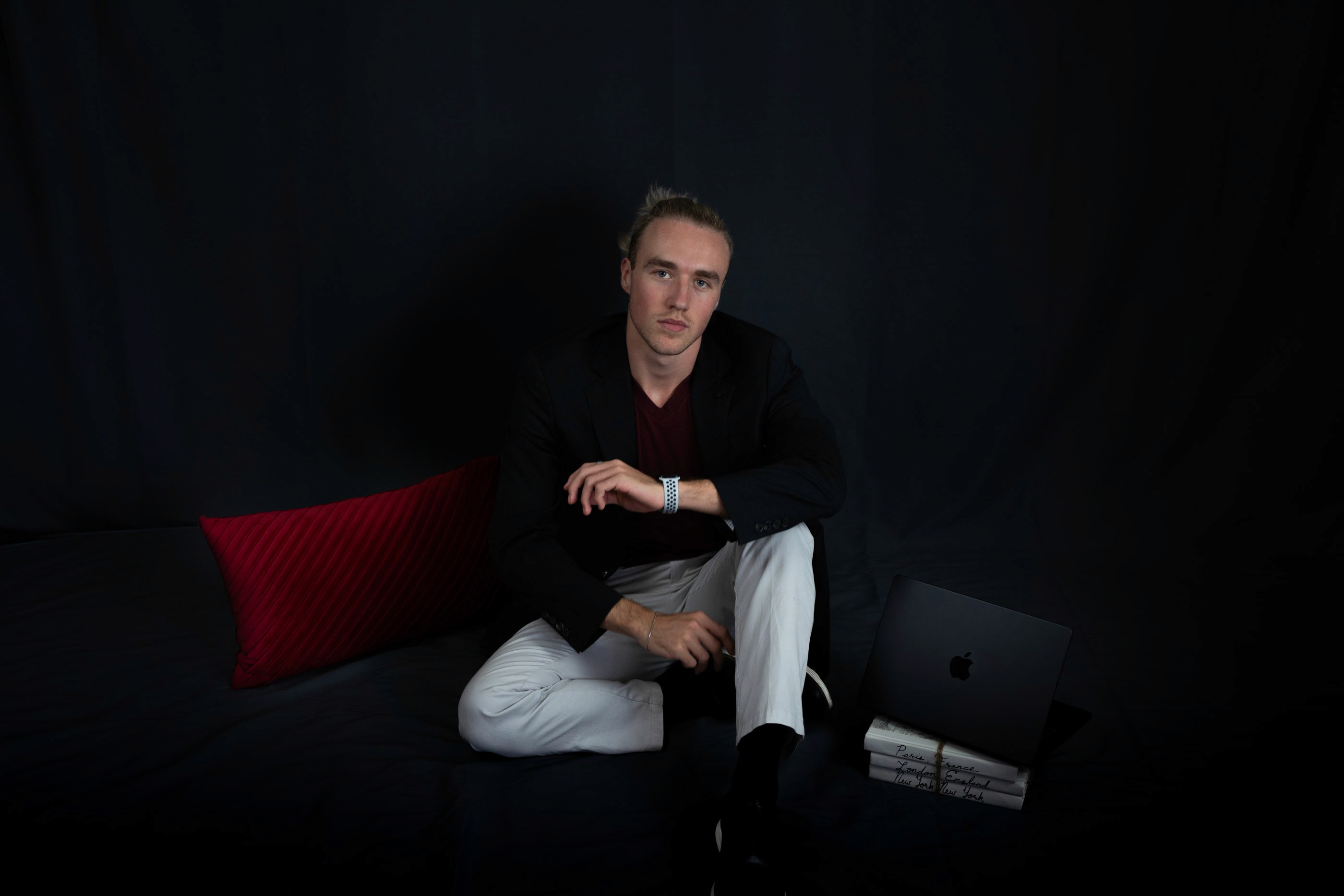A man sitting in front of a red pillow with a MacBook Pro sitting next to him
