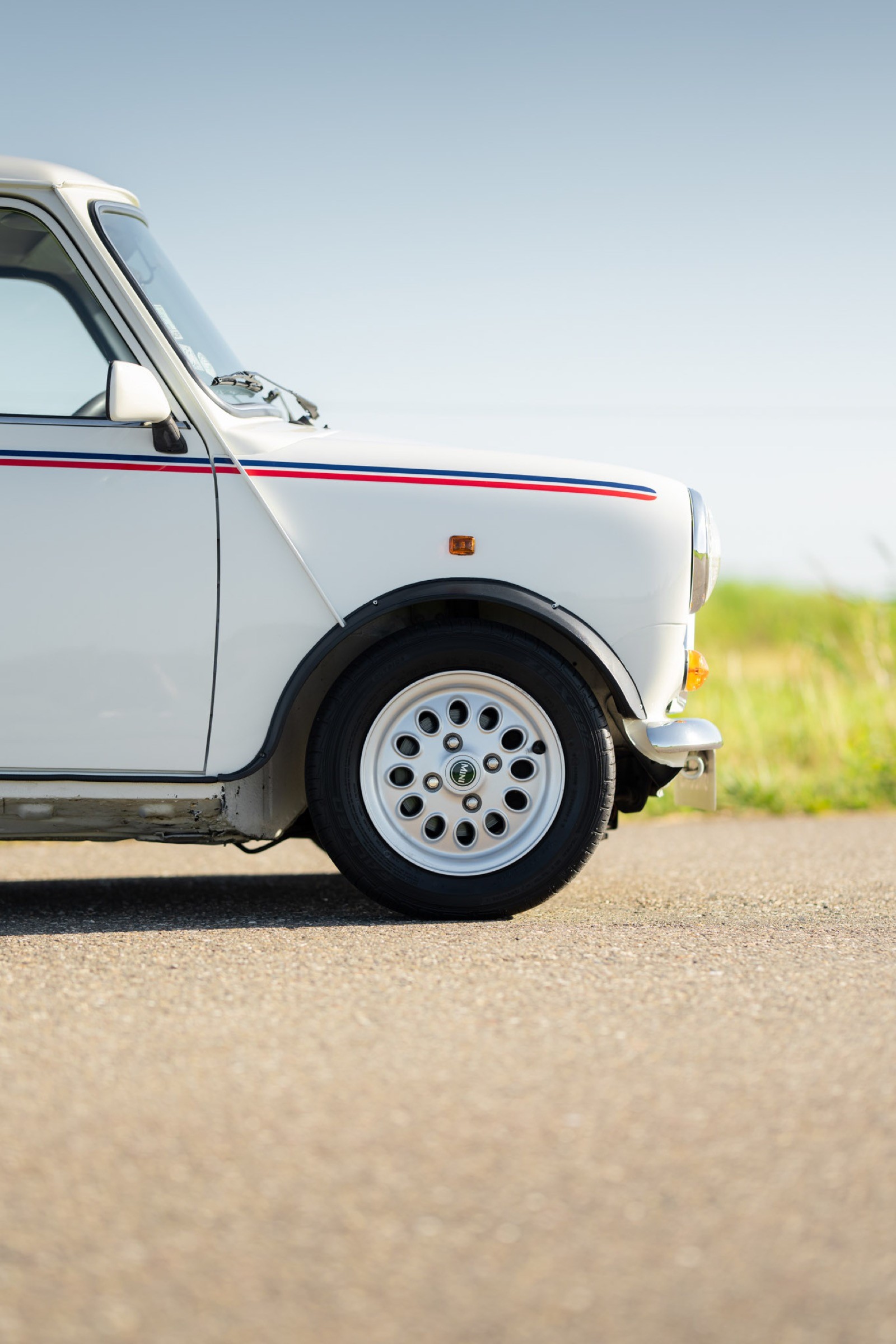 A close-up of a white vintage car parked on a road, with green grass and clear sky in the background.
