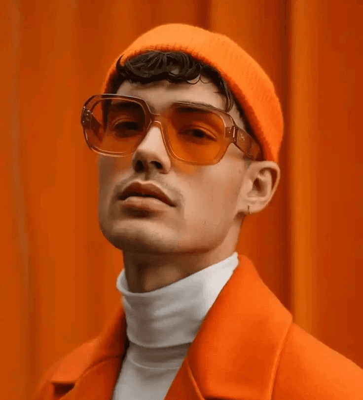 A person wears an orange beanie, matching coat, and tinted sunglasses over a white turtleneck. The rich orange background creates a bold, cohesive look.
