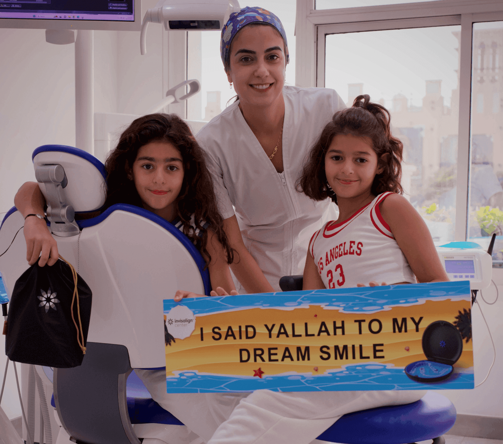 طبيبة أسنان أطفال بمركز إنفزلاين الطبي مع طفلتان يبتسمون للكاميرا و تمسك إحدى الطفلتين بلافتة تعبر عن سعادتها بابتسامتها الصحية والجميلة.