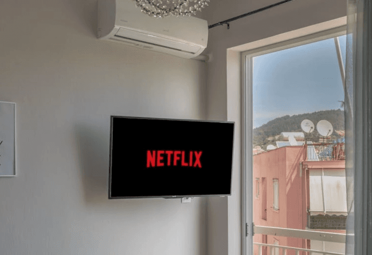 실내 벽면에 설치된 TV 화면 가득 넷플릭스 로고가 떠 있는 모습입니다. 창밖으로 산과 다른 건물들이 보이는 원룸 공간의 인테리어와 홈 시네마 환경 상징합니다.