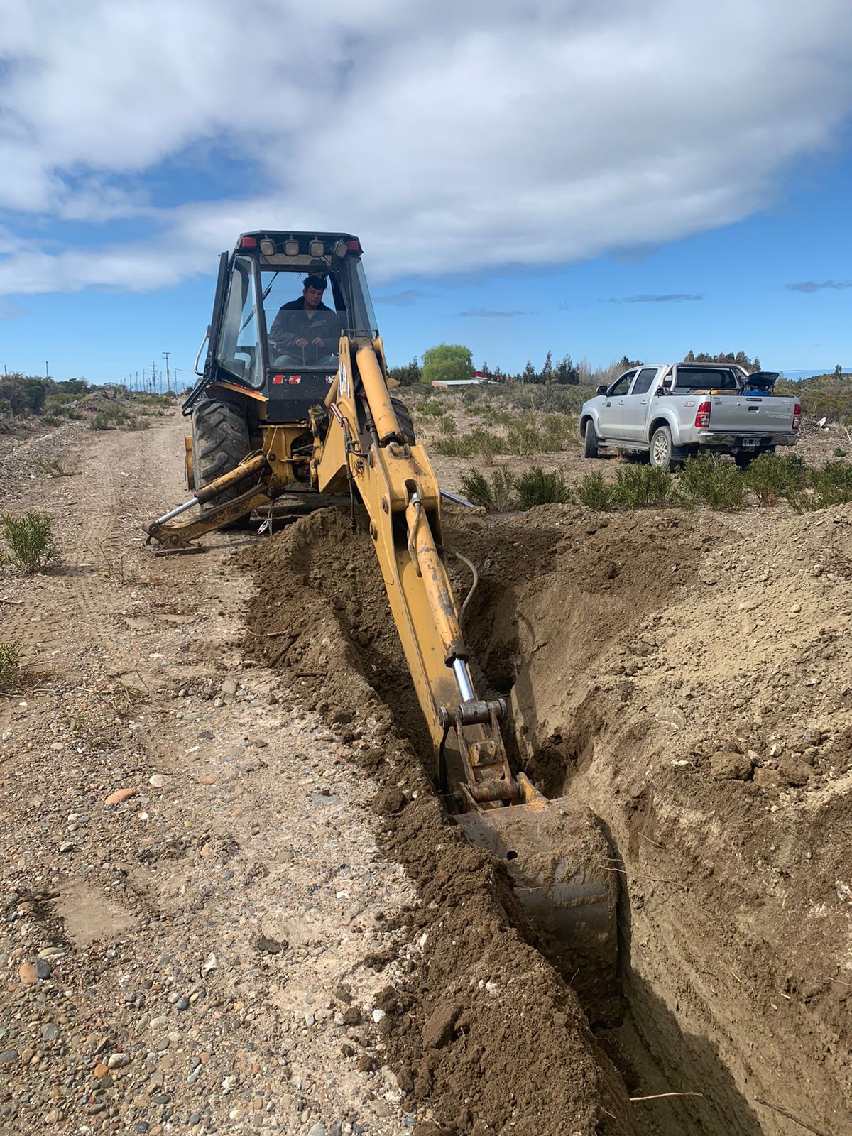 A yellow excavator digging a trench in an open area under a blue sky with scattered clouds.