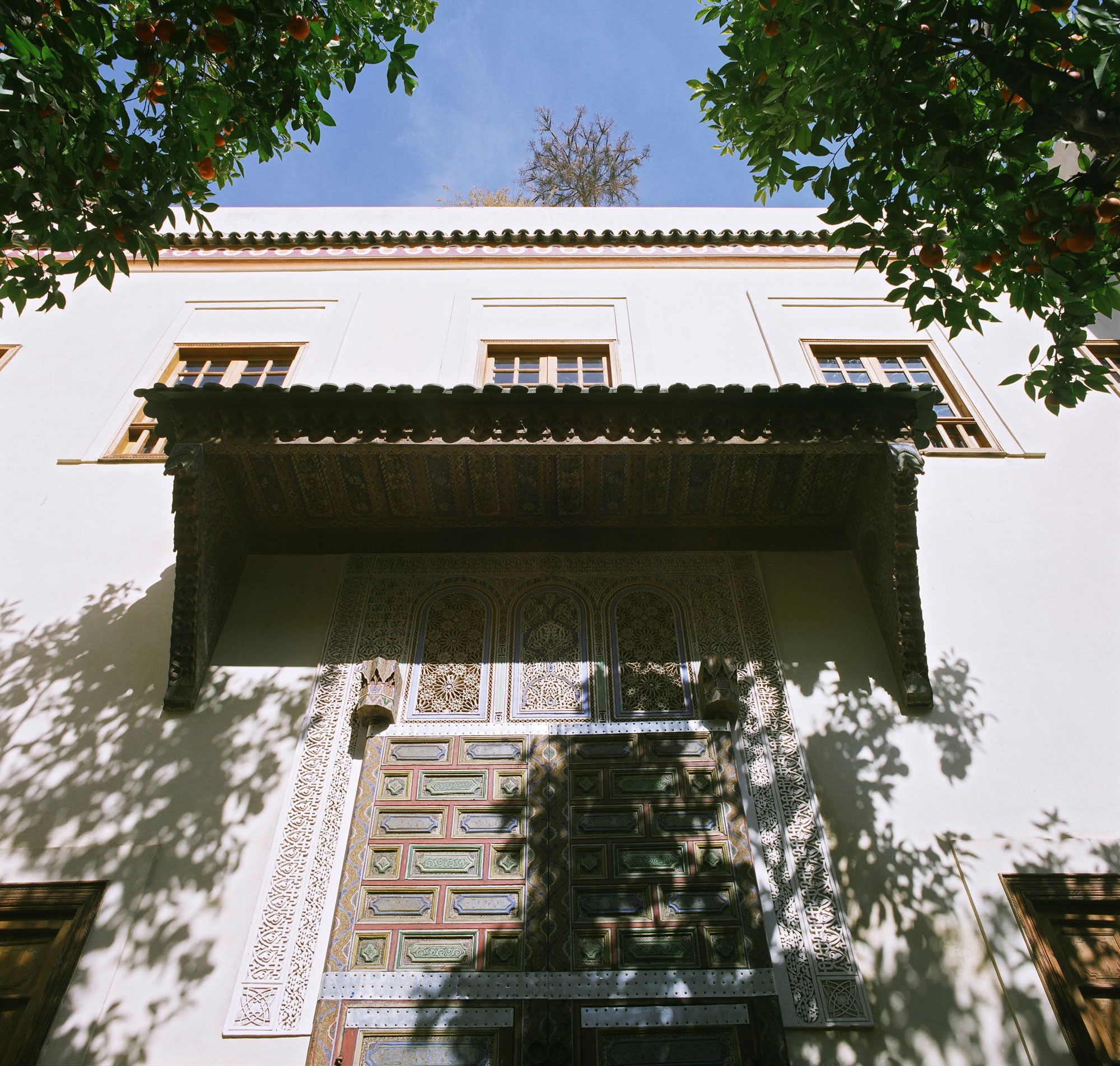 Facade with ornate wooden gate and decorative details, tree branches casting shadows on the sunlit wall.