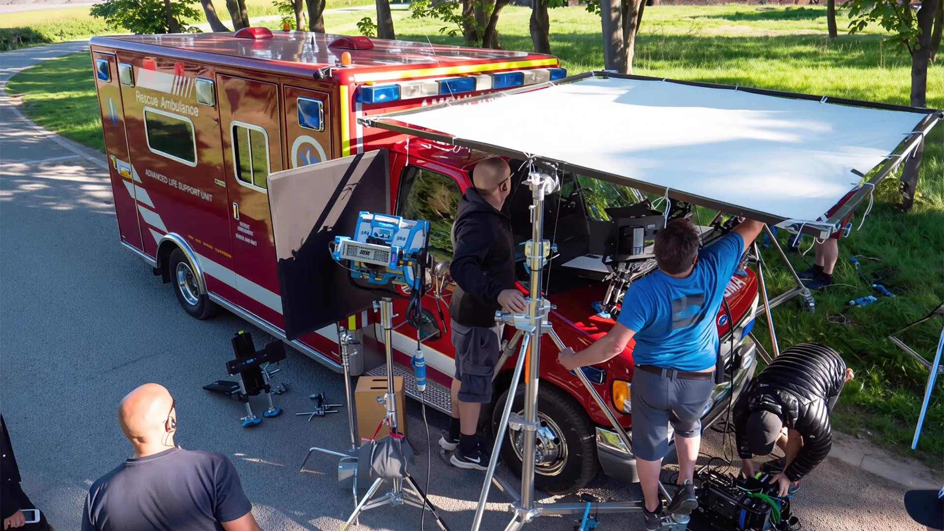 Film crew setting up lighting and camera equipment beside a rescue ambulance on a roadside
