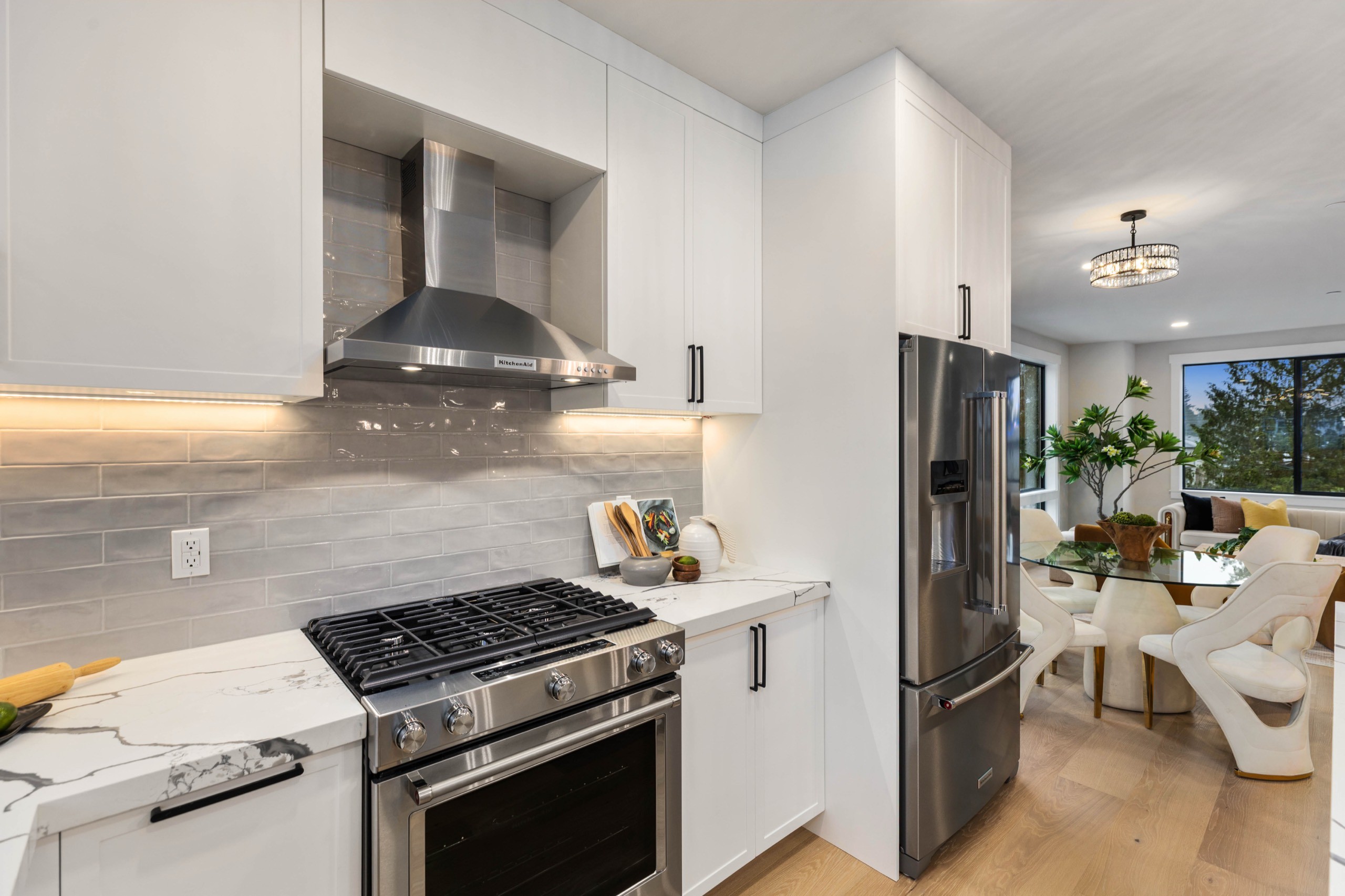 Compact luxury kitchen with white cabinetry, grey marble tile backsplash, stainless steel gas range, wall-mounted range hood, and open dining area with chandelier