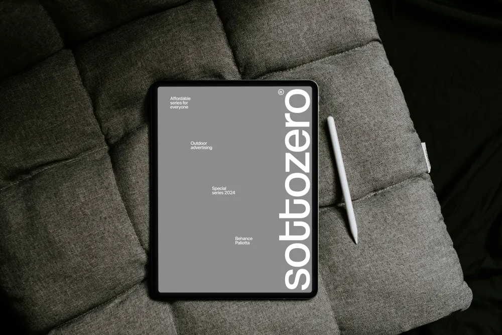 iPad mockup placed on a textured gray cushion, accompanied by a white stylus. The screen displays minimalist branding with a vertical logo and text elements. Cozy indoor setting.