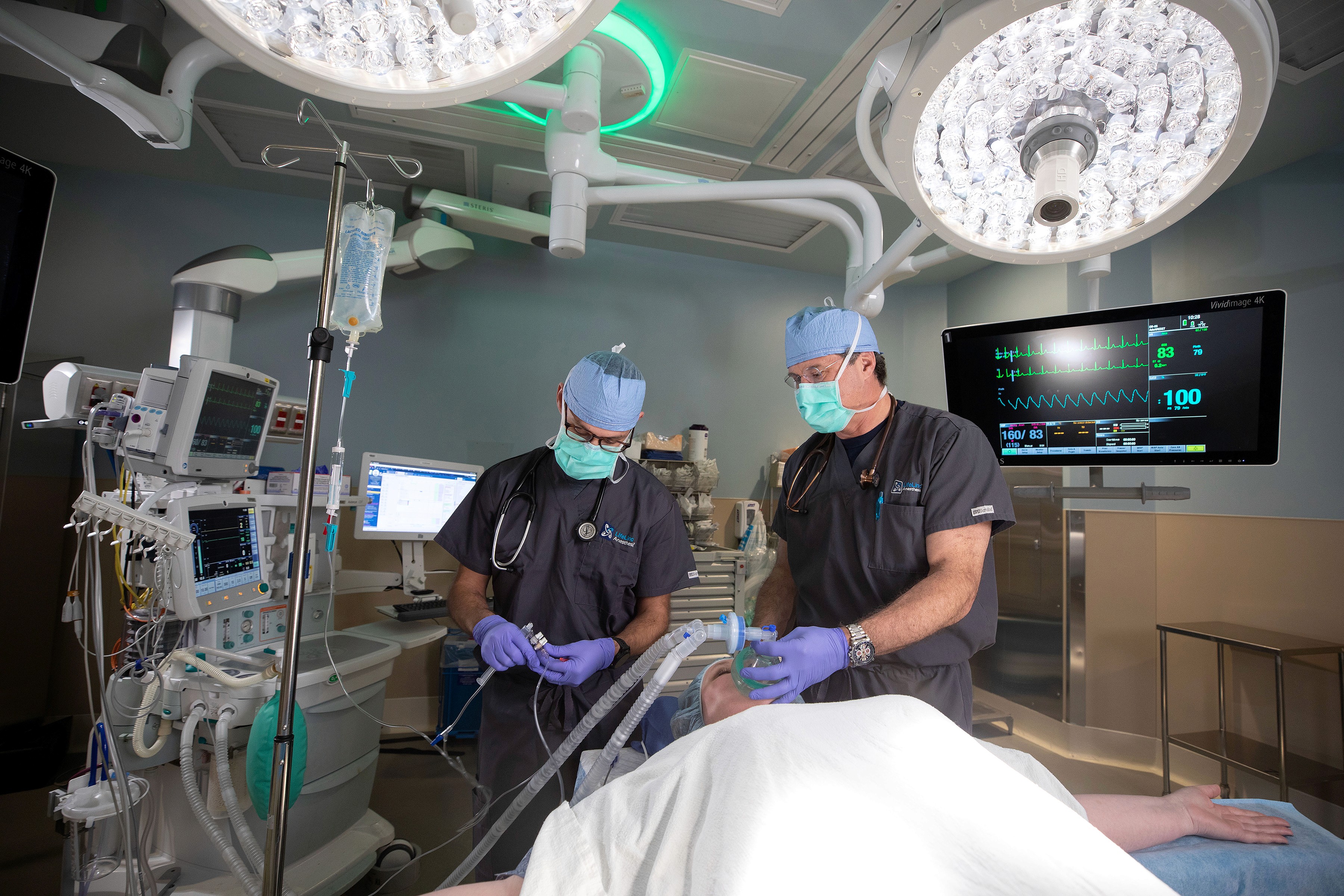 An anesthesia care team prepares a patient for surgery in an ambulatory surgery center operating room, with overhead surgical lights and monitoring equipment visible