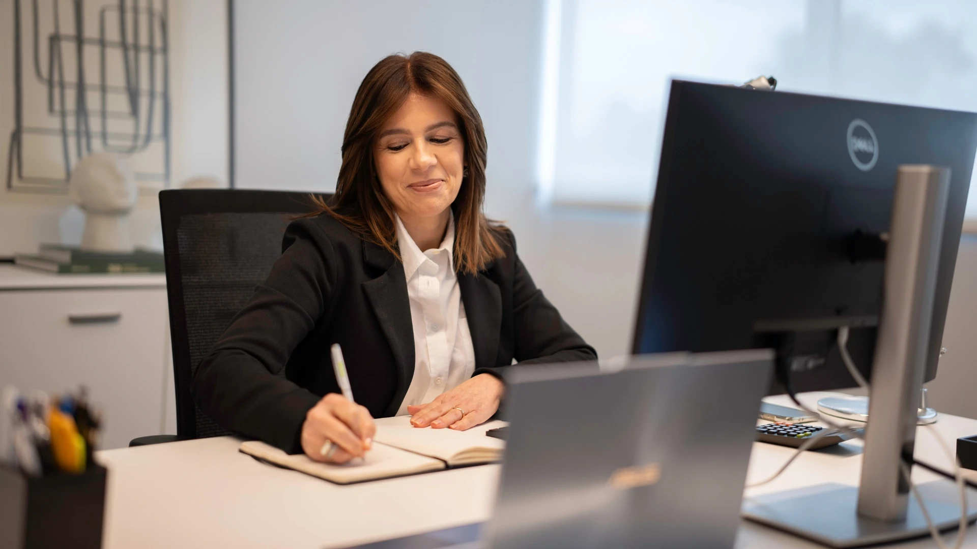 Mulher executiva sorrindo enquanto trabalha em escritório com computador e caderno, evidenciando ambiente corporativo profissional.