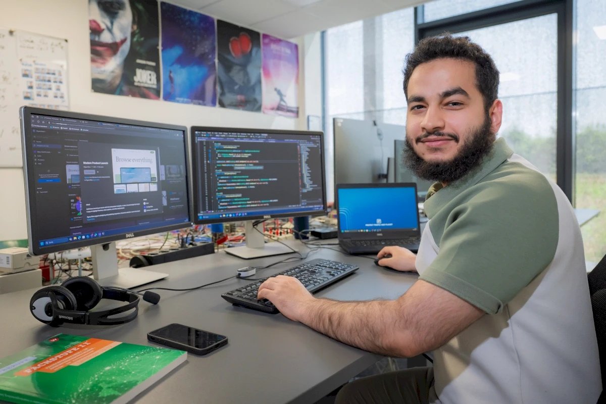 Mann lächelt in die Kamera, während er an einem Computer-Setup arbeitet, auf dem Code auf den Monitoren angezeigt wird.