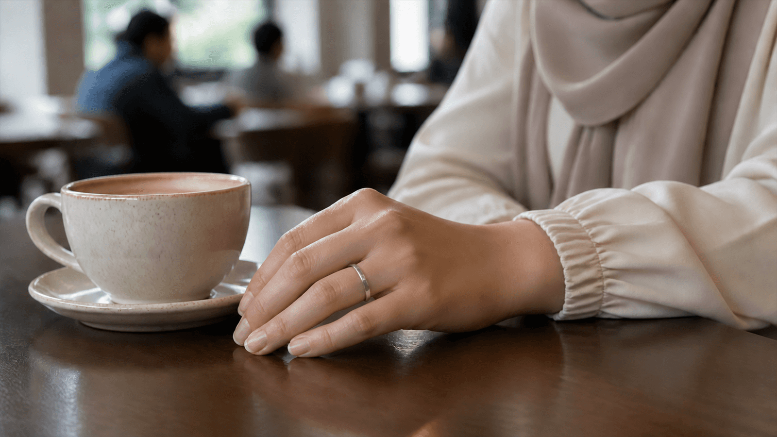 A woman's hand resting on a cafe table mid-commute, a zikr ring worn like everyday jewelry