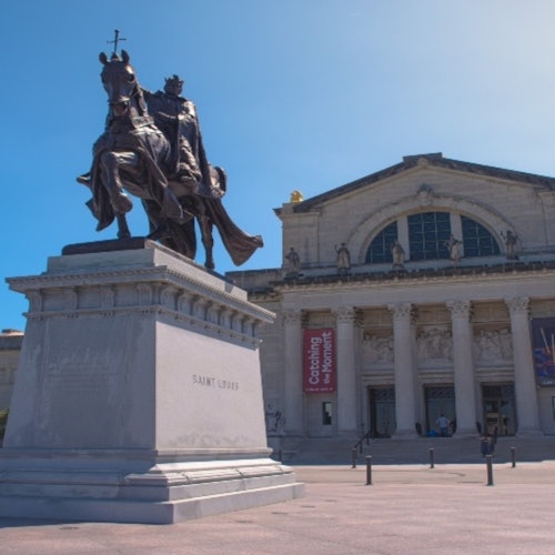 Equestrian statue of Saint Louis in front of a classical building with large columns and banners. Clear blue sky above.