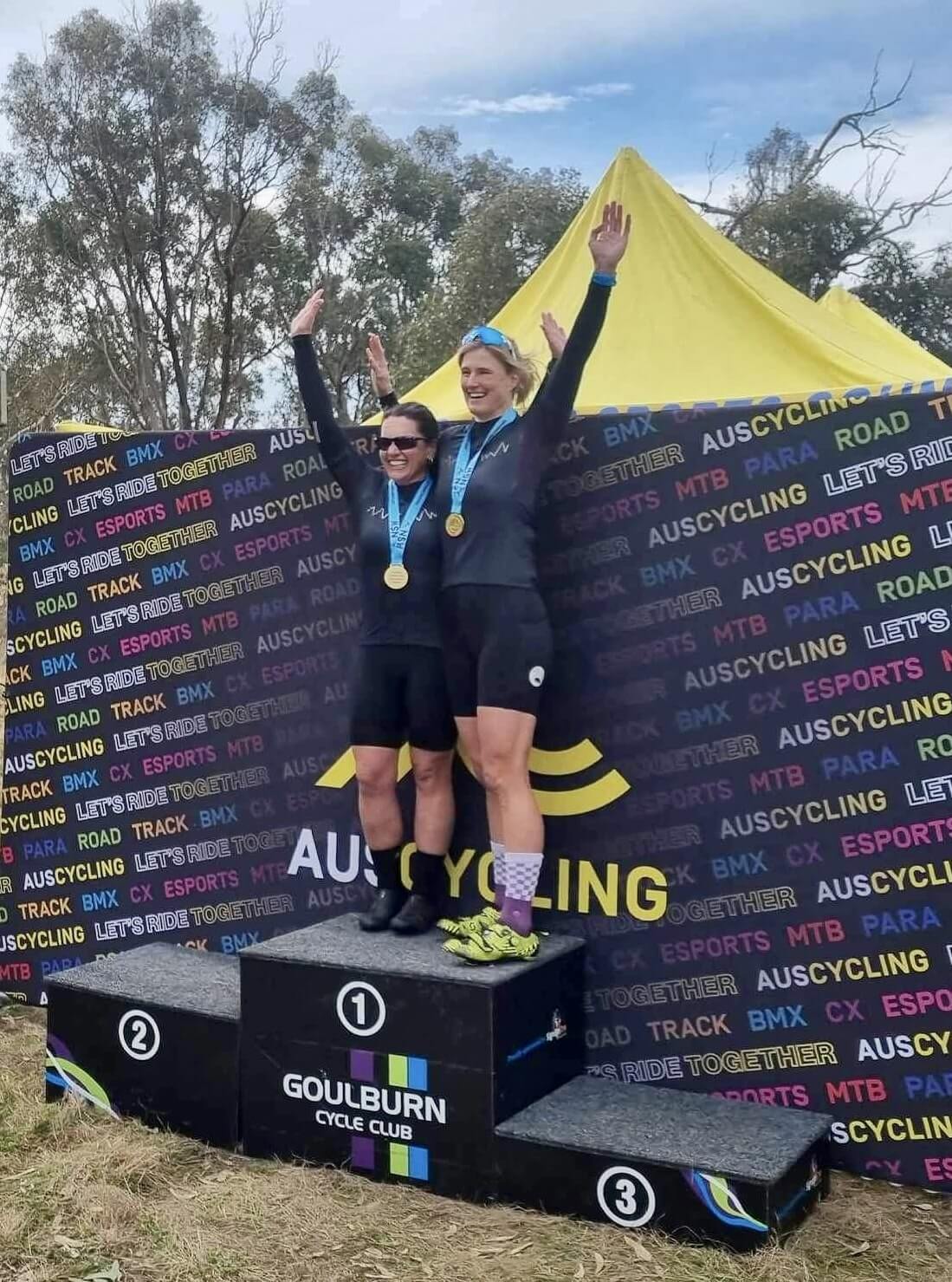 Lily and Jay posing triumphantly on a first place podium with their gold medals