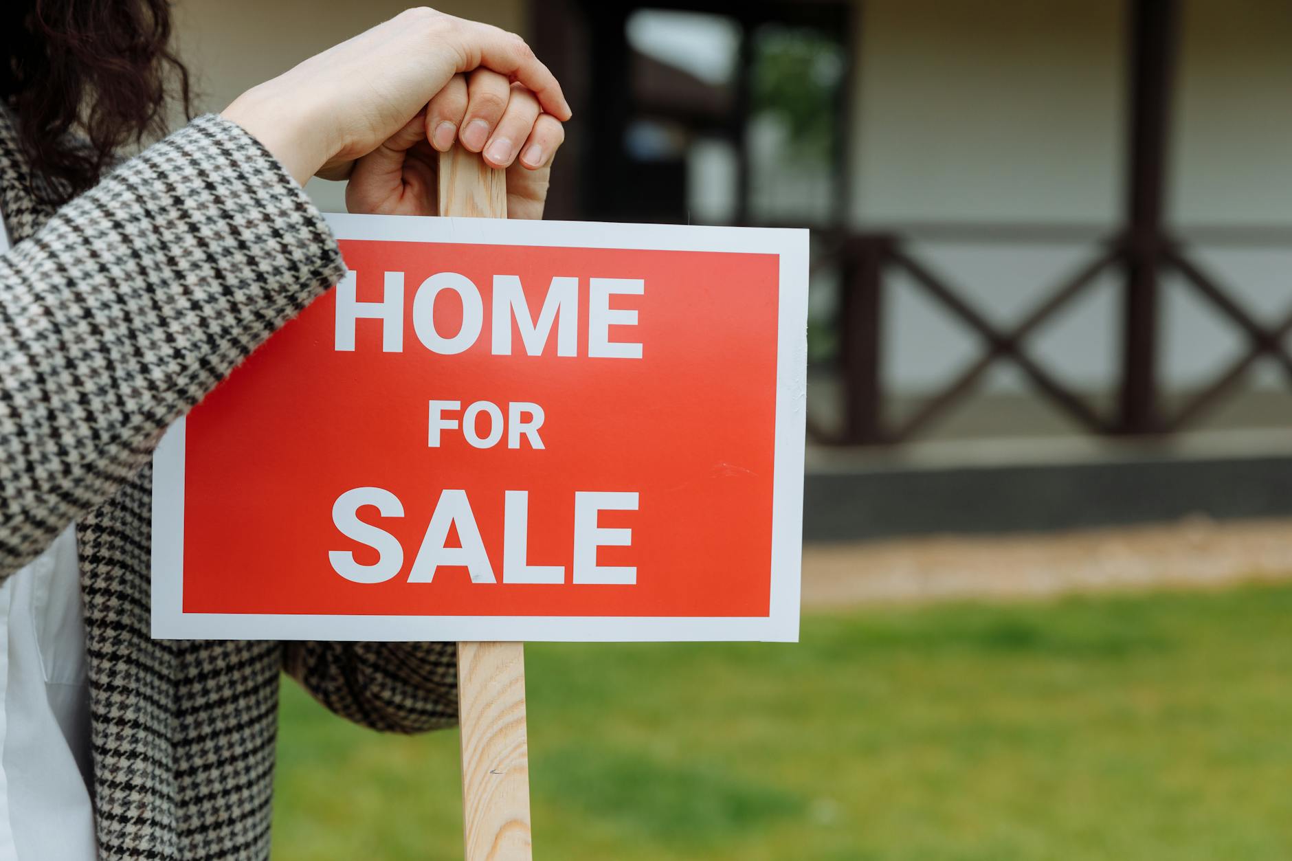 Woman in a houndstooth jacket holding a red Home For Sale sign in front of a white house with wooden porch railings
