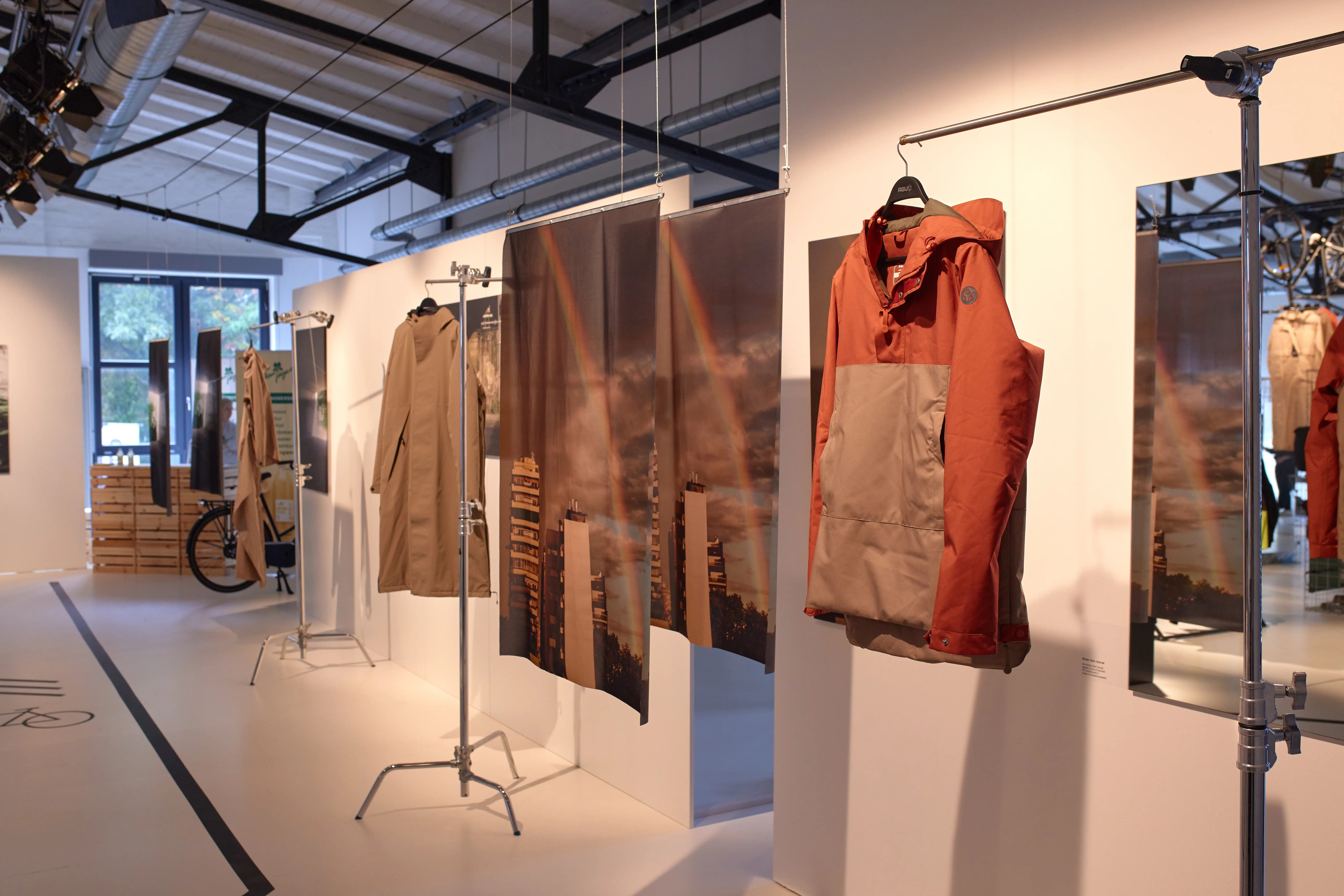 Exhibition aisle at the AGU showcase with rain jackets and large urban photo prints hanging from steel stands.
