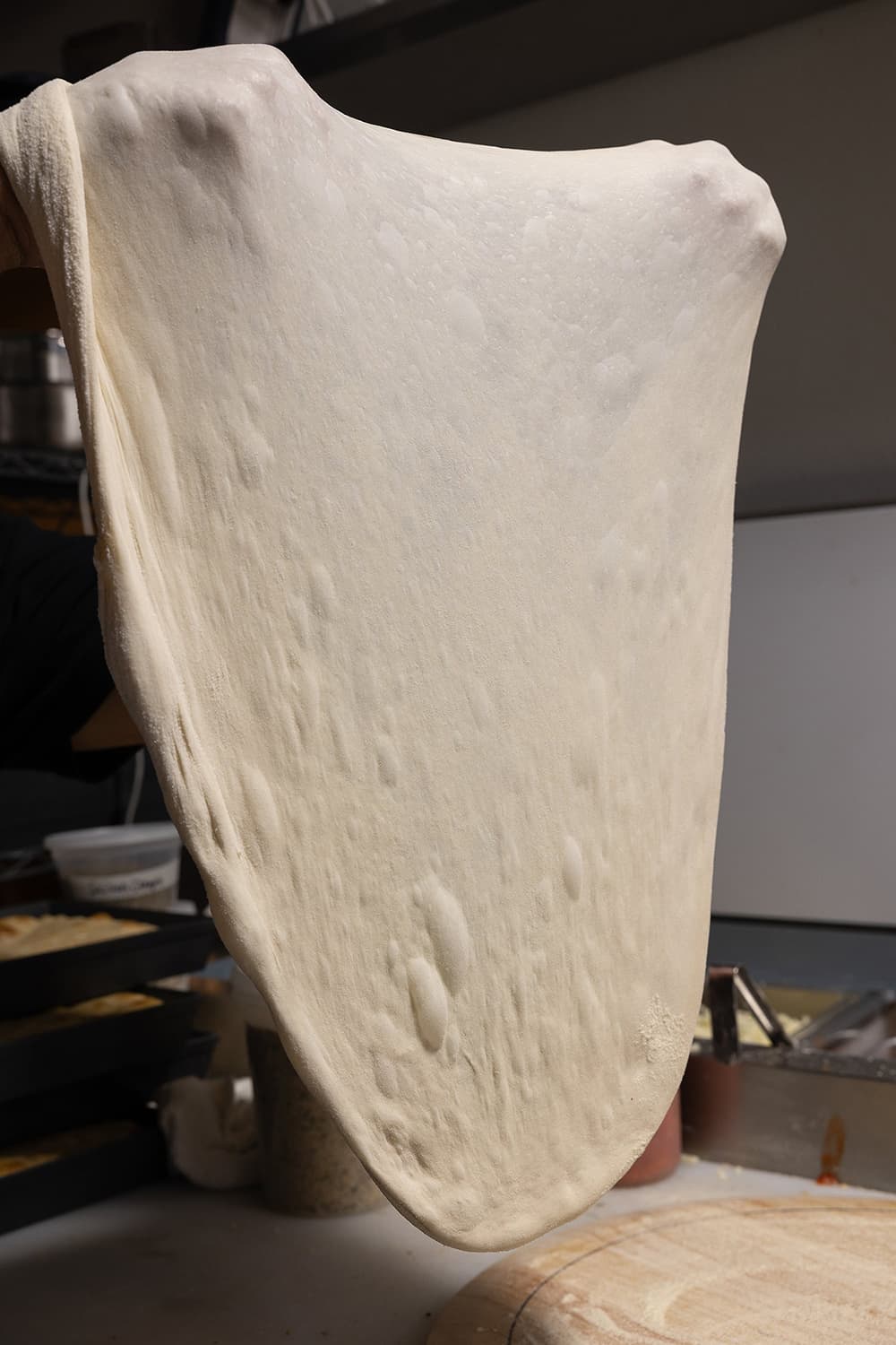 Pizza dough being stretched thin over a work surface, showing air bubbles and an elastic texture in a kitchen setting.