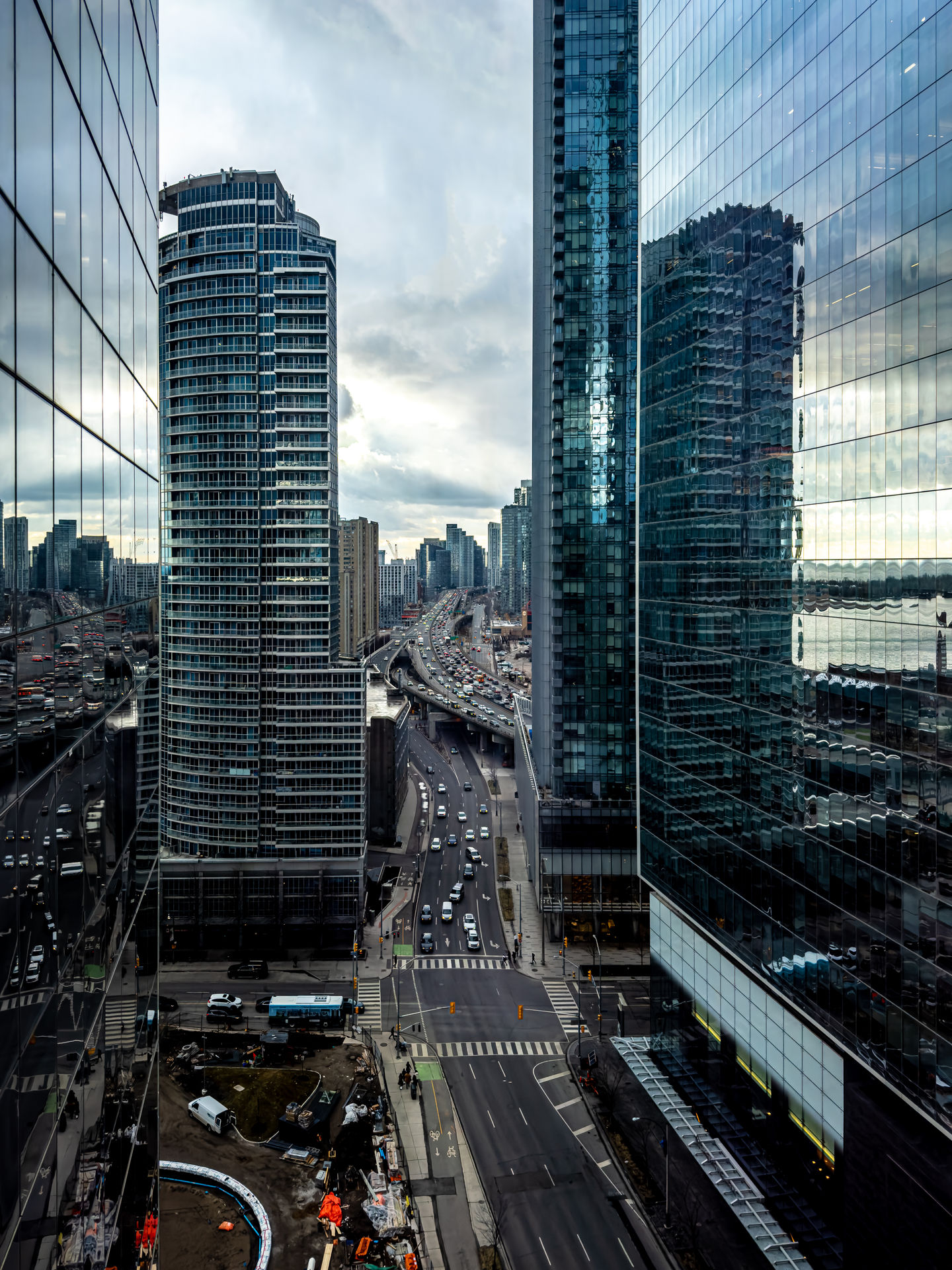 Urban view of a busy multilane highway cutting through a downtown core, surrounded by modern high-rise buildings with reflective glass exteriors. Cars fill the road below under an overcast sky, while a construction site occupies a corner in the lower left of the frame.