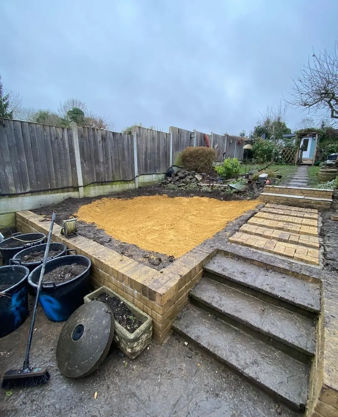 A construction site with wooden steps leading to a raised area filled with yellow sand and surrounded by fences.