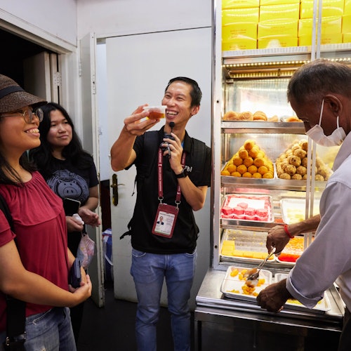A man holding a sweet while speaking, with three people watching near a display case filled with various sweets inside a store.